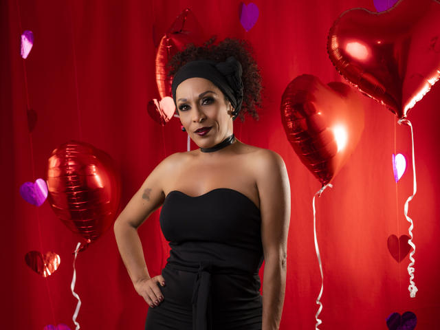KhazandraX with curly hair, black headband, choker, strapless black dress, standing, hands on hips, red balloons, hearts, and purple hearts, dark lipstick, confident smile.