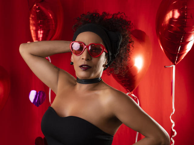 KhazandraX with curly hair, black headwrap, red heart glasses, black choker, and strapless black top, hand behind head, red balloons, dark lipstick, looking at camera.