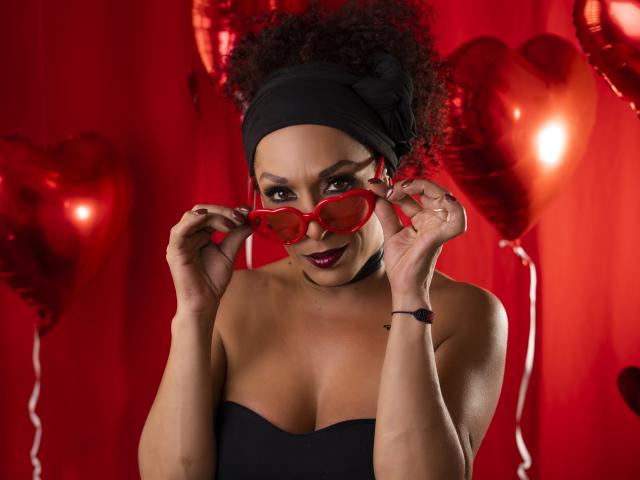 KhazandraX with curly black hair, red heart-shaped glasses, black headband, and strapless black top, holding glasses, smiling, showing medium brown tits, red background, heart balloons.