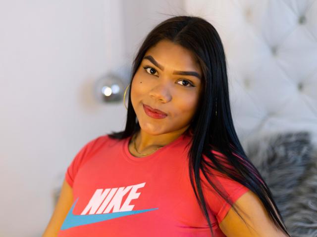 NickyBernad with long black hair, brown eyes, red lipstick, and hoop earrings, wearing a red Nike shirt, smiling softly, has a small beauty mark on her right cheek.