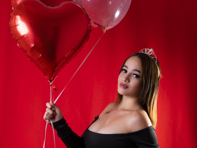 LuciRaven mit langen, braunen Haaren, trägt ein schwarzes Oberteil mit einem Schulterauschnitt, hält rote Herzballons und einen pinken Ballon, trägt eine silberne Tiara, hat kleine Brüste, roter Hintergrund..