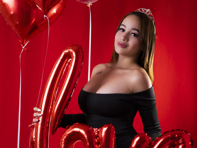 LuciRaven mit großen Brüsten, trägt ein schwarzes, schulterfreies Kleid, hält rote Herzballons, hat ein süßes Lächeln und trägt eine silberne Tiara, vor einem roten Hintergrund..