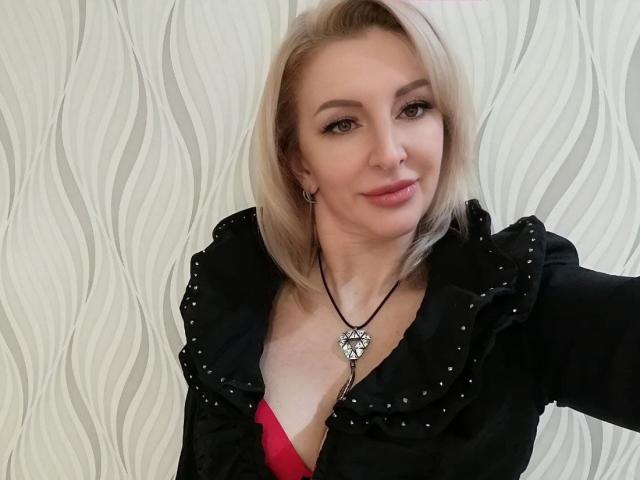 RoseQMagic with blonde hair, wearing a black shirt with studs, red bra visible, necklace with flower pendant, smiling, light skin, subtle makeup, and a nose ring.
