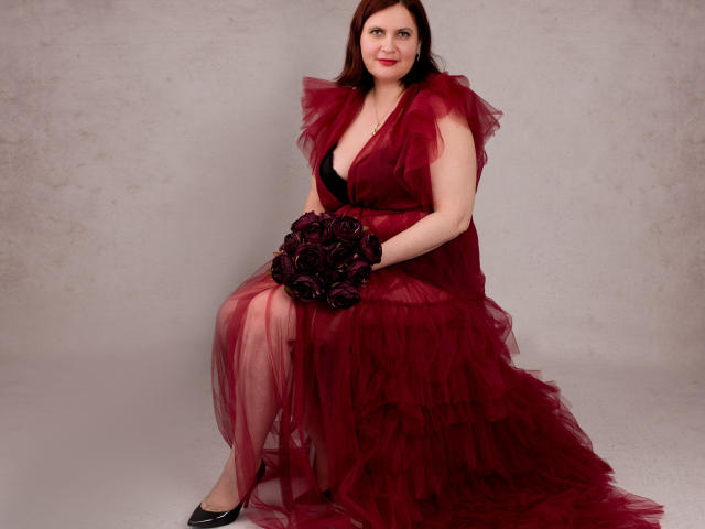 BigTitsXHot with red, sheer, ruffled dress, black gloves, and bouquet of red roses, sitting, holding flowers, wearing black heels, pale skin, dark red hair, smiling.