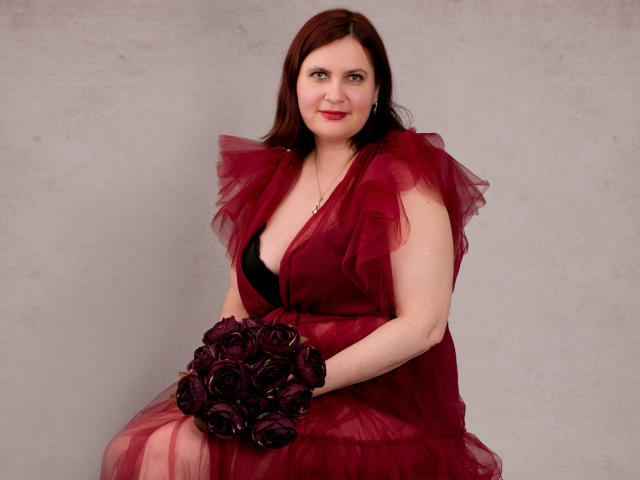BigTitsXHot met een rode tule jurk, zwarte bh, een boeket paarse rozen vasthoudend, zittend, lachend, bleke huid, rode lippenstift, halflang bruin haar, grijze achtergrond..