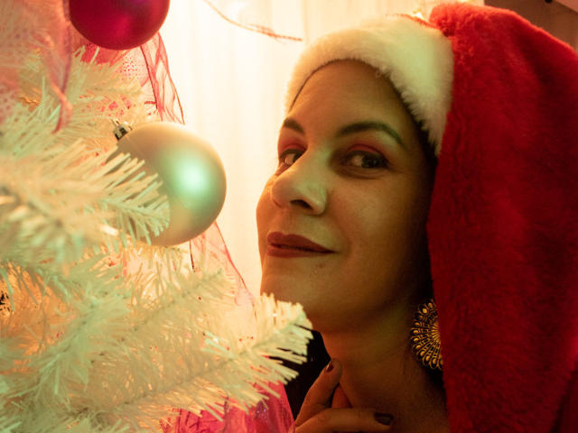 RoraimAnaggie with a red Santa hat, smiling at a white Christmas tree with blue and pink ornaments, her hand on her neck, wearing a gold earring, in warm light.