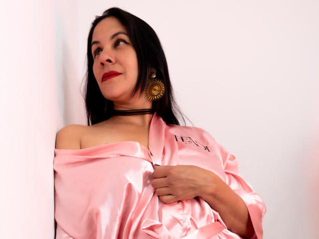 RoraimAnaggie, con cabello negro largo, lápiz labial rojo y aretes dorados en forma de sol, vestida con una bata de satén rosa, con la mano en el pecho, mirando hacia arriba con una ligera sonrisa..