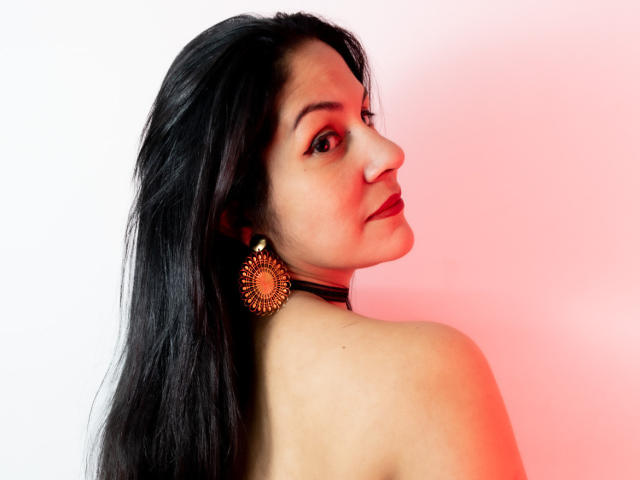 RoraimAnaggie with long black hair, brown eyes, and red lipstick, wearing a black choker and gold sunburst earrings, looking over her shoulder, bare shoulders, red light.