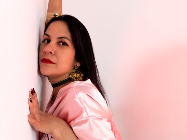 RoraimAnaggie with long black hair, red lipstick, and gold earrings, wearing a pink satin shirt, leans against a white wall, arm raised, hand with black nails touching wall.