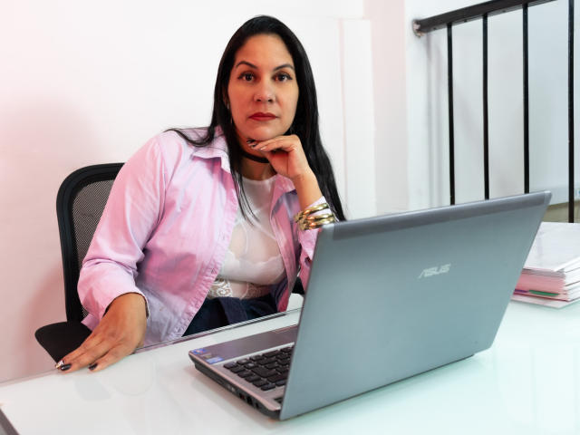 RoraimAnaggie, met lang zwart haar, een roze shirt en een witte blouse, zit aan een bureau, met haar hand op haar kin, en kijkt naar een ASUS laptop. Ze draagt gouden armbanden..