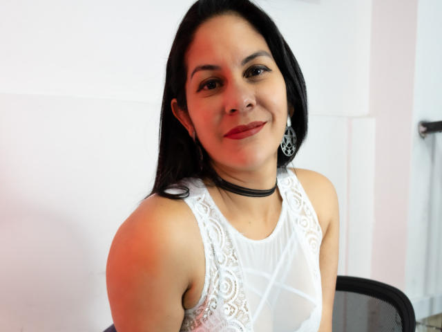 RoraimAnaggie has dark straight hair, red lipstick, white lace top, black choker, and silver earrings, smiling slightly at the camera. She's sitting with a black chair behind her.