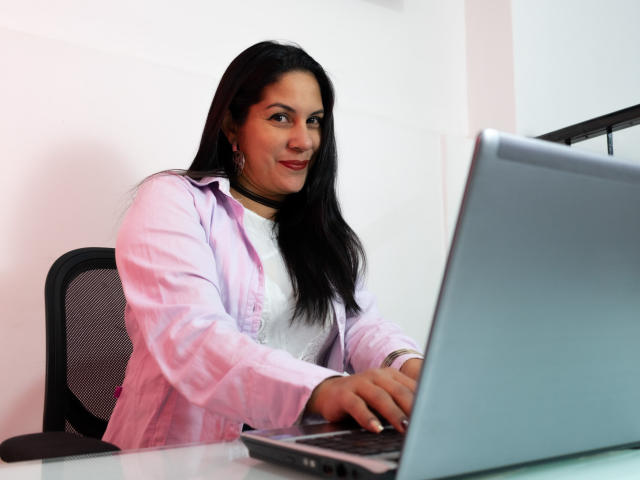 RoraimAnaggie with long black hair, pink shirt, white top, and black chair, typing on a laptop, showing a cute smile, small tits, and a confident, sexy look.