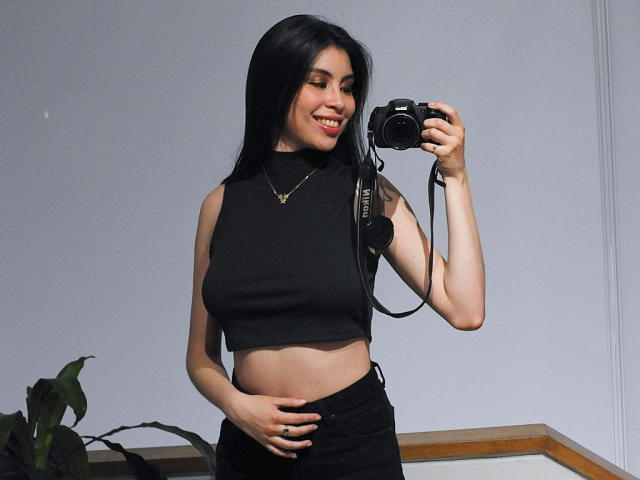 IsaTimeSweet mit langen, schwarzen Haaren, trägt ein schwarzes, ärmelloses Crop-Top und schwarze Hosen, hält eine Kamera, lächelt und berührt ihren Bauch. Ihre Brüste sind unter dem Top sichtbar..