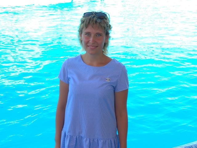DianaBritt stands in front of a bright blue pool, wearing a light purple dress with a small emblem on the chest, and sunglasses on her head, smiling.