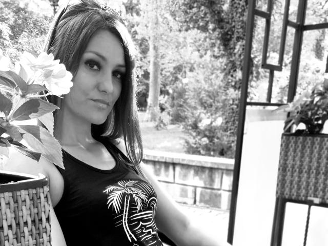 Black-and-white photo of JoliDanielle with long, straight hair, wearing a black tank top with a white leaf design, showing her small tits and a slight smile.