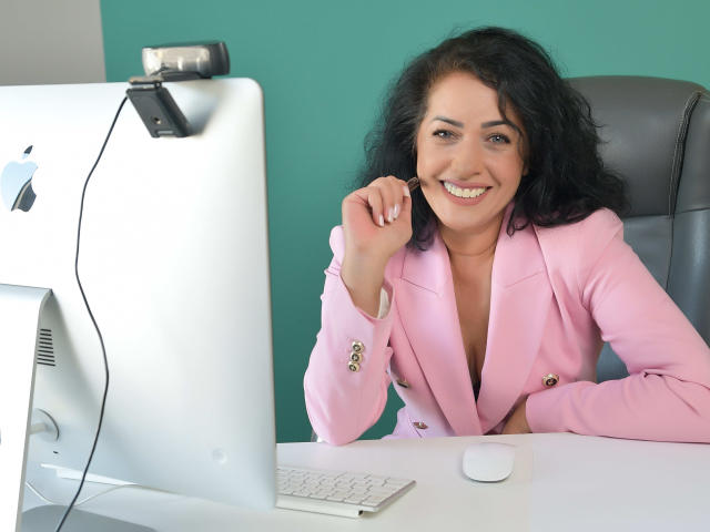 AnelisaReys, avec ses cheveux noirs bouclés, souriante, portant un blazer rose, est assise à un bureau blanc avec un Apple iMac, un clavier blanc et une souris, et une caméra au-dessus..