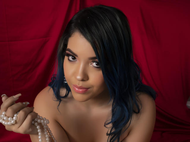 KimLafayette with dark blue hair, holding pearl necklace, bare shoulders, sexy brown eyes, slight smile, red background, soft makeup, medium skin, subtle lipstick, looking up at camera.