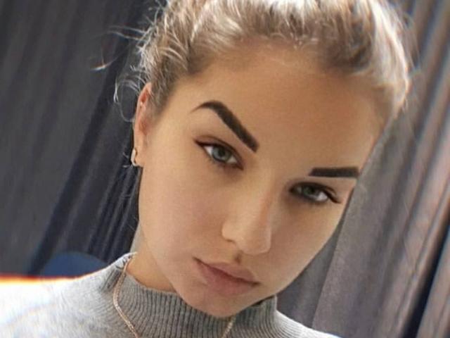 barbiandken, mit hochgezogenen Augenbrauen, grünen Augen und dezentem Make-up, trägt einen grauen Rollkragenpullover, hat ein kleines Nasenpiercing und hellbraunes Haar, das zu einem unordentlichen Dutt gebunden ist..
