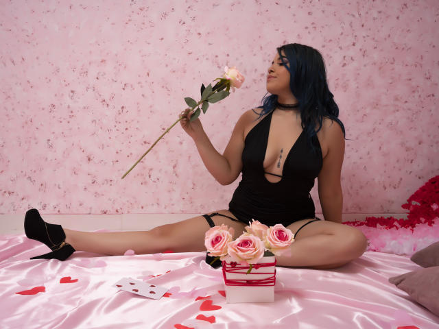 KimLafayette with blue hair, black halter dress, garter, black heels, holding rose, sitting on pink satin, roses in front, tattoo on chest, legs spread, sexy.