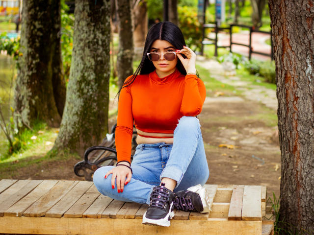 BrithneyLee, avec ses longs cheveux noirs, portant un haut court orange, un jean bleu et des baskets noires, est assise en tailleur sur un banc en bois, et ajuste ses lunettes de soleil..