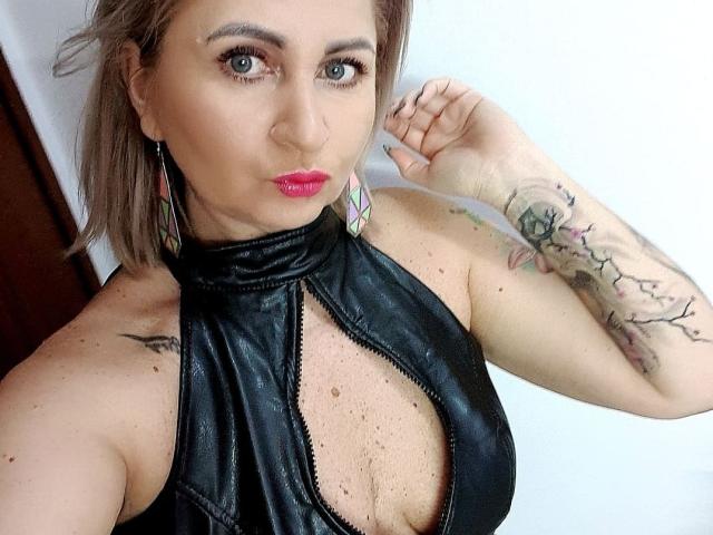 ChaudeEvely with short brown hair, wearing a black leather halter top, exposing her cleavage, has tattoos on her right arm, and is pouting with red lipstick.
