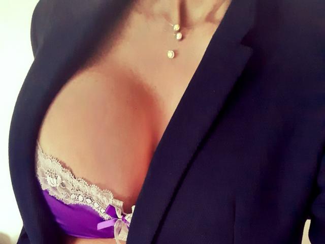 Il seno ampio e scoperto di LaParisienne, con un reggiseno di pizzo viola con bordi in pizzo bianco, a malapena coperto da un blazer nero, che mostra una collana d'oro con piccole perle rotonde pendenti..