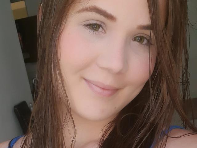 Emily Suarez tiene el cabello castaño y húmedo, ojos verdes y una ligera sonrisa con labios rosados, viste una blusa azul, mostrando una piel suave y pálida, y una pequeña marca de nacimiento cerca de su boca..
