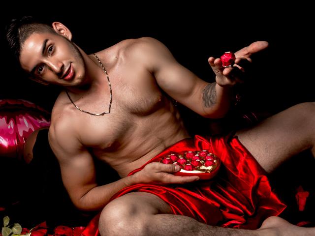 JonathanJeremiah, shirtless with red satin shorts, holding a rose and a plate of strawberries, has a tattoo on his left arm and a chain necklace, looking seductive.