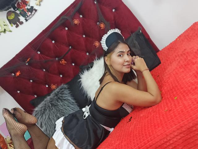 ArianaGuzman69 deitada em uma cama, vestindo um uniforme de empregada preto e branco, meias de rede, e uma faixa de cabeça preta, com um cobertor vermelho, olhando para a câmera..