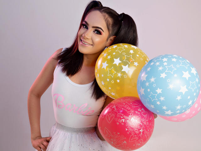 LunaRuiiz, com maria-chiquinhas, blusa branca e saia brilhante, segurando balões rosa, amarelos e azuis com estampas de estrelas, sorrindo com os seios levemente visíveis sob a blusa..