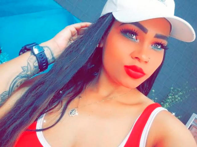 LinaaPrincess, com cabelos longos e pretos, usando um boné branco, uma blusa vermelha e branca, maquiagem azul nos olhos, batom vermelho, uma tatuagem no pulso e um relógio preto, olhando para a câmera..