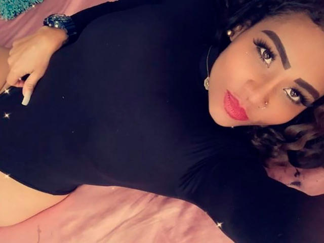 LinaaPrincess lying on a pink bed, wearing a black long-sleeve leotard, showing her tits, with red lipstick, a nose ring, and a bracelet, looking at the camera.