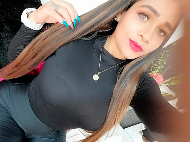 LinaaPrincess, com cabelos castanhos longos, blusa preta, unhas azuis, batom rosa, colar de ouro e relógio branco, exibindo seus seios e traseiro curvilíneo..
