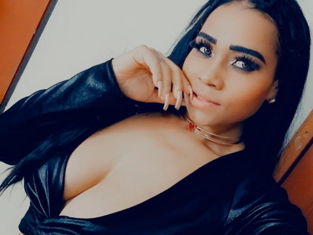 LinaaPrincess has big tits, long black hair, and thick eyebrows. She's wearing a black leather top, a gold choker with a red gem, and is touching her mouth.