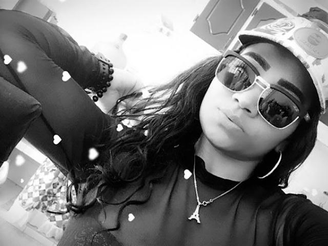 LinaaPrincess, com cabelos longos e escuros, usando um boné, óculos de sol, brincos de argola e um colar, tem uma expressão de lábios franzidos e veste um cachecol de gola alta, com o braço levantado para uma selfie..
