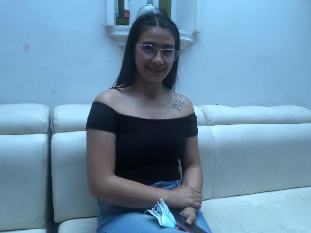 JadeNielsen with glasses, black off-shoulder top, blue jeans, holding mask, tattoo on left shoulder, sitting on white leather couch, smiling.