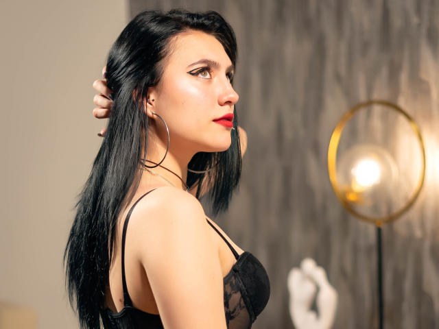 MolySmith heeft zwart haar, rode lippenstift en draagt een zwarte kanten BH, waardoor haar borsten zichtbaar zijn, met grote oorbellen. Ze staat in profiel, met haar hand in haar haar, onder een lamp..