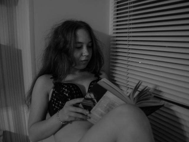 AnikaGlow with messy hair, wearing a black sequined bra, sitting, reading a book, showing her tits, in a dimly lit room with blinds. Monochrome, relaxed.