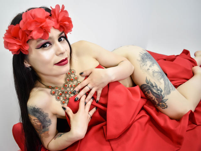 JolieDeluxe avec une coiffe en forme de fleur rouge, une robe rouge et des bijoux, allongée sur un tissu rouge, cachant ses seins avec ses mains, des tatouages sur les bras, du vernis à ongles noir, du rouge à lèvres rouge, et souriante..