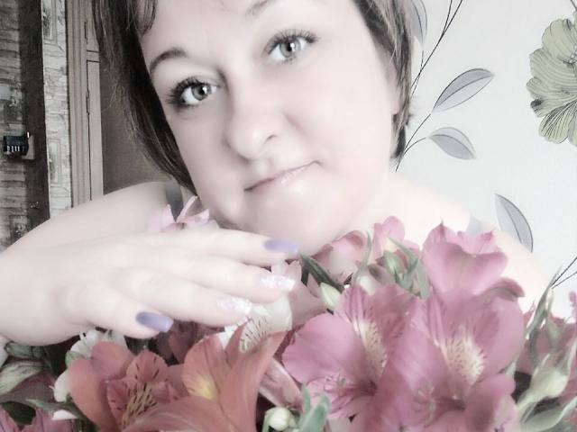 NikaMalay with short brown hair, fair skin, and purple nails, holding pink lilies, smiling softly, eyes slightly squinted, wearing a floral dress, looking cute and sweet.