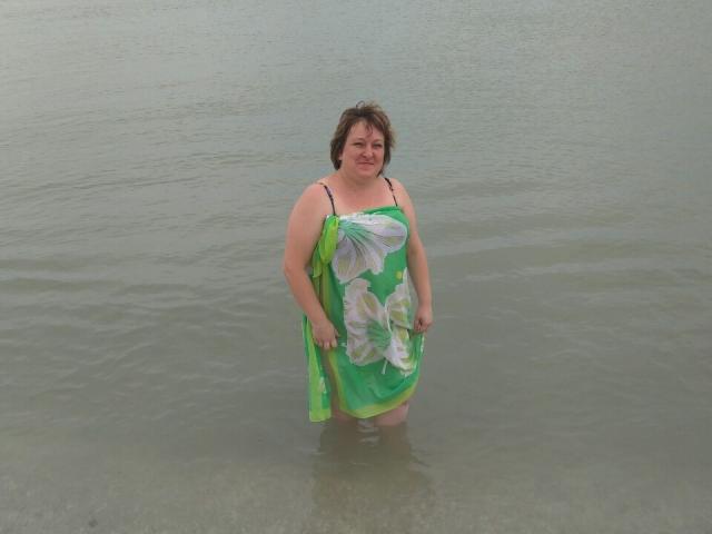 NikaMalay has short brown hair, a chubby build, and wears a green floral dress standing in shallow water, with her tits visible through the wet fabric.