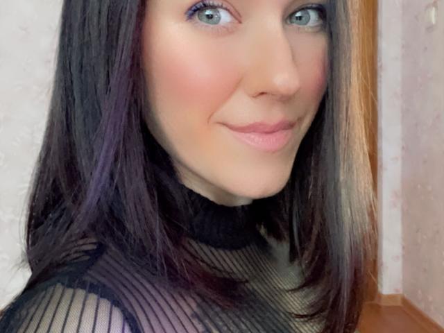 Misstyk with blue eyes, pale skin, and pink lips, wearing a sheer black top, has dark brown hair with purple highlights, smiling softly at the camera.