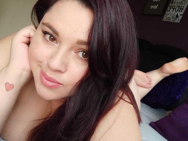 LissaClaire with dark brown hair, pink lips, and a small heart tattoo on her left wrist, lying on a bed, smiling at the camera, legs up in the background.