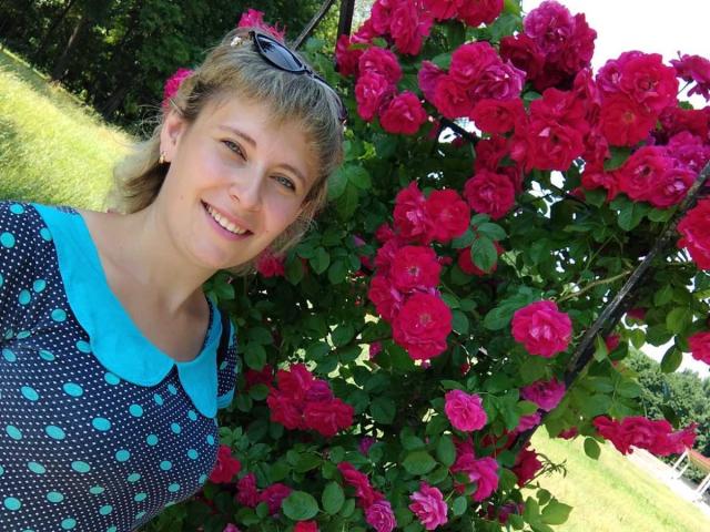 DianaBritt, con i capelli biondi, indossa un abito blu a pois con colletto turchese, e sorride accanto a un cespuglio di rose rosa acceso. Ha gli occhiali da sole in testa e sullo sfondo si vedono piante di colore verde..