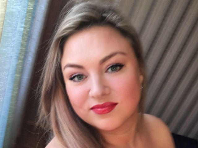 HugeNaturalTits ha i capelli biondi, gli occhi verdi e il rossetto rosso. Sorride con un trucco leggero, indossando una maglietta nera. Il suo seno non è visibile. È illuminata da una luce soffusa..