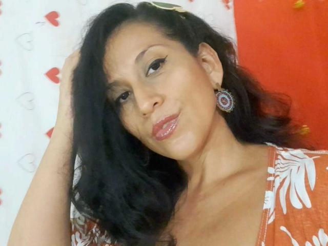 KattyBoobsLove, con i capelli scuri e ondulati, indossa una maglietta arancione a fiori, ha labbra rosa lucide e sta toccandosi la testa, guardando leggermente di lato, indossando un orecchino viola..