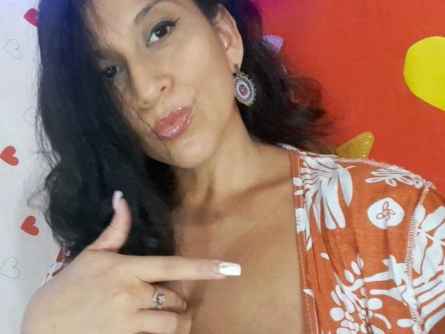 KattyBoobsLove, met een gemiddelde borstmaat, bruine huid en zwart golvend haar, draagt een oranje top met bloemenprint en wijst met haar witte nagels naar haar borst. Ze heeft glanzende, roze lippen en rode oorbellen..