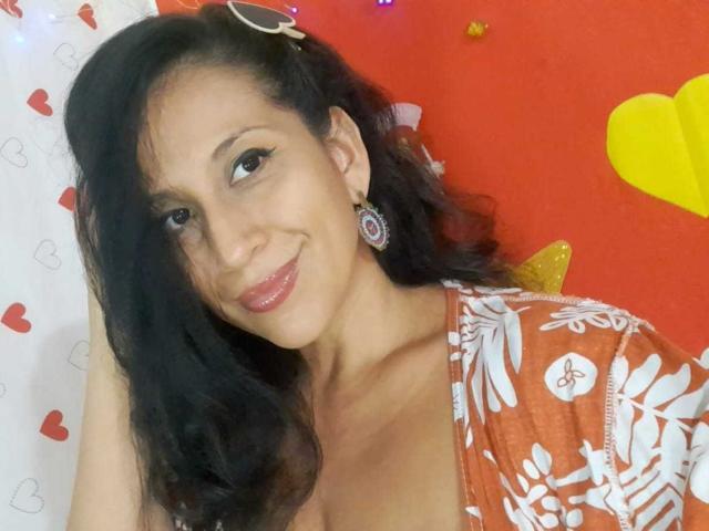 KattyBoobsLove, con cabello negro largo, vistiendo una blusa roja con estampado floral, sonriendo, mostrando su escote, usando aretes y un clip para el cabello, en un selfie, luciendo linda y sexy..