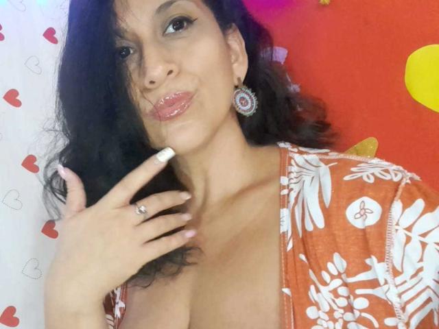KattyBoobsLove, avec ses cheveux noirs ondulés, ses lèvres roses brillantes, portant un haut orange à motifs floraux, dévoilant sa poitrine, et touchant son menton avec un doigt orné d'une bague, portant des boucles d'oreilles ornées..