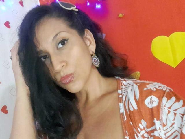 KattyBoobsLove con cabello oscuro y ondulado, labios rosados brillantes, vestido naranja con estampado floral blanco, aretes dorados y una expresión de labios fruncidos, mirando a la cámara. Fondo rojo con un corazón amarillo y luces de cadena..